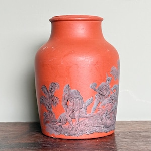 May include: A terracotta-colored vase with a detailed relief design. The relief depicts figures, dogs, and foliage in a monochromatic gray. The vase has a rounded body and a slightly flared neck, showcasing a classic aesthetic.