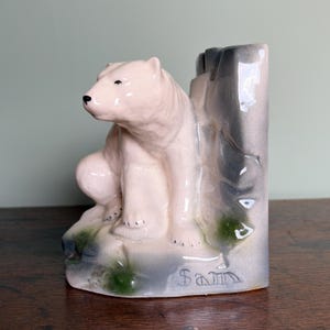 Könnte beinhalten: Eine Keramikfigur eines Eisbären in Weiß, Grau und Grün, sitzend neben einer stilisierten Felsformation. Auf der Basis der Figur ist der Name "Sam" eingraviert. Die Figur steht auf einer Holzoberfläche.