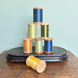 May include: A pyramid stack of six wooden spools wrapped in dark blue, olive green, light green, and orange thread, with one additional yellow thread spool resting horizontally in front on a dark oval base with a worn finish.