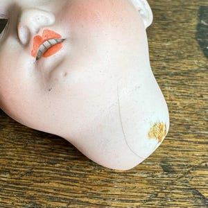 May include: A vintage doll head with a pale complexion, rosy cheeks, and orange-red lips. The porcelain head shows signs of age, with a crack and discoloration on the chin. The doll's closed eyes and serene expression evoke a sense of nostalgia.