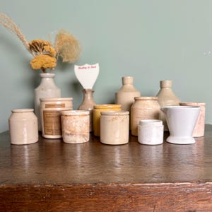 May include: A collection of 10 small, cream-colored ceramic jars and vases, some with a brown glaze, on a wooden table.