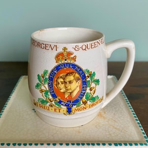 May include: Cream-colored ceramic mug with a handle, featuring a royal crest with portraits and text. The mug sits on a square, off-white plate with a turquoise border. The mug has the words "GEORGE VI & QUEEN ELIZABETH" on it.
