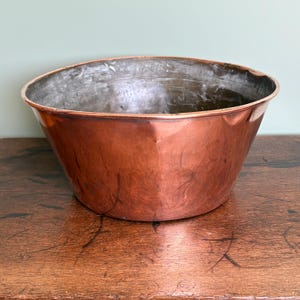 May include: A large, antique copper bowl with a wide, open top and a gently sloping, faceted body. The bowl has a rich, reddish-brown patina, and the interior shows signs of age and use. The bowl sits on a dark wooden surface.