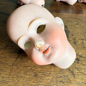 May include: A vintage porcelain doll head with painted features. The doll head has a pale complexion, rosy cheeks, and painted orange lips. The eyebrows are arched and painted gold. The doll head is lying on a wooden surface.