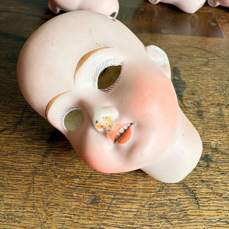 May include: A vintage porcelain doll head with painted features. The doll head has a pale complexion, rosy cheeks, and painted orange lips. The eyebrows are arched and painted gold. The doll head is lying on a wooden surface.