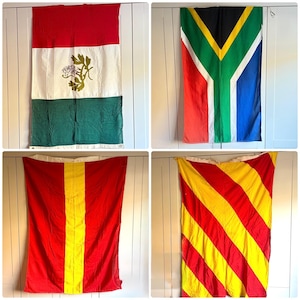 May include: Four colorful flags hanging on a white wall. The flags include the Mexican flag, the South African flag, a red flag with a yellow vertical stripe, and a red and yellow striped flag.