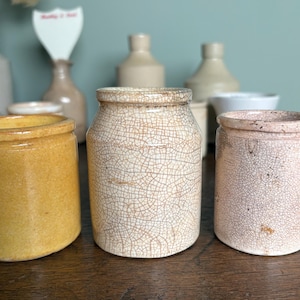 May include: A collection of vintage ceramic jars with crackle glaze. The jars are in various shades of white, yellow, and pink. The jars are displayed on a wooden surface.
