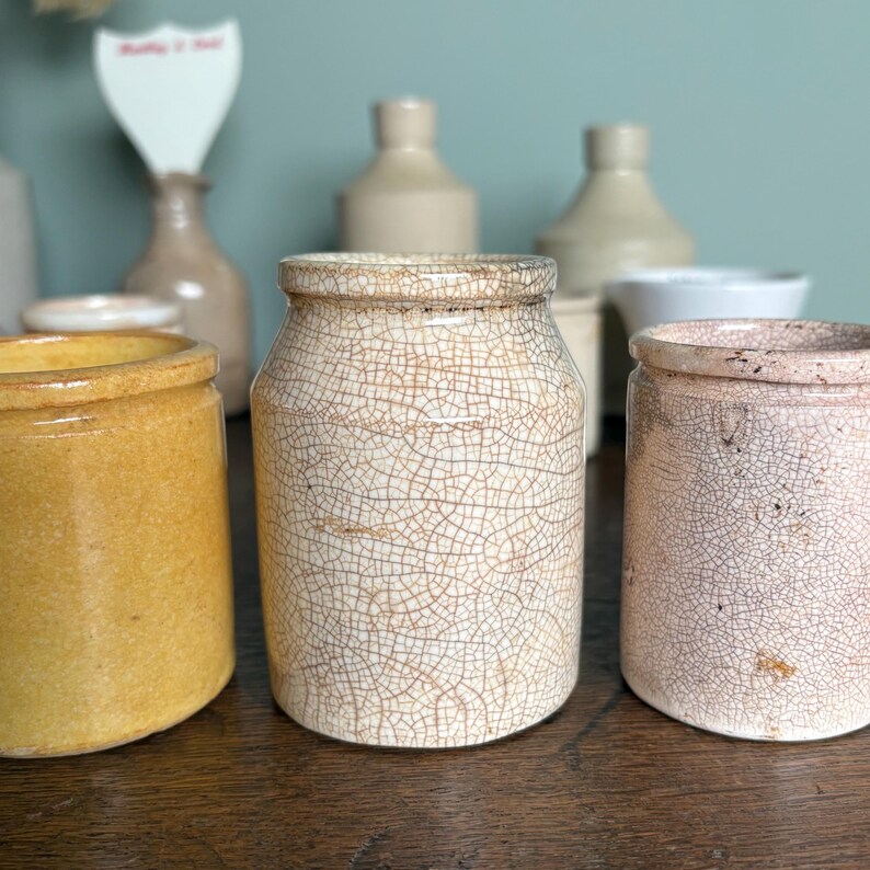 May include: A collection of vintage ceramic jars with crackle glaze. The jars are in various shades of white, yellow, and pink. The jars are displayed on a wooden surface.