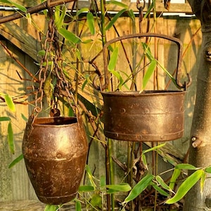 Könnte beinhalten: Zwei antike, rostige Metall-Hängetöpfe mit brauner Oberfläche. Ein Topf wird an einer Kette aufgehängt, der andere hat einen Griff. Der Hintergrund zeigt Bambus und einen Holzzaun, was auf eine Außendekoration hindeutet.