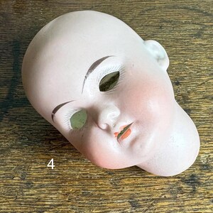 May include: A vintage porcelain doll head with painted facial features. The head is pale pink with rosy cheeks, brown eyebrows, and orange lips. The eye sockets are open, and the number "4" is visible.