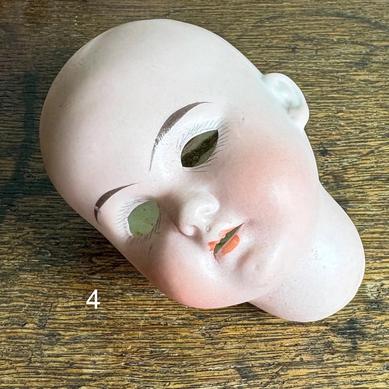 May include: A vintage porcelain doll head with painted facial features. The head is pale pink with rosy cheeks, brown eyebrows, and orange lips. The eye sockets are open, and the number "4" is visible.