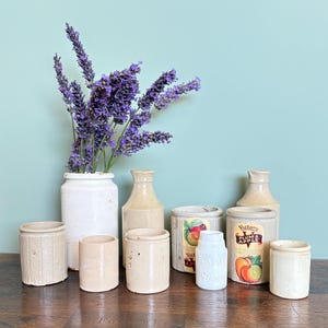 May include: A collection of antique stoneware jars and bottles, some with labels, arranged on a wooden surface. A white jar holds a bouquet of purple lavender. The jars vary in size and shape, with a rustic, aged appearance.