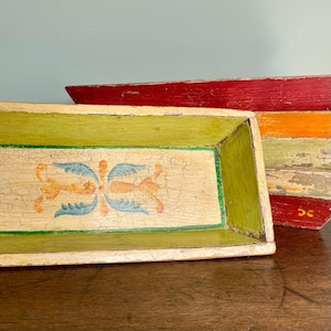 Op de afbeelding: Twee houten trays met beschilderde ontwerpen. De bovenste tray is rood, oranje en geel. De onderste tray is groen, geel en blauw met een bloemmotief.