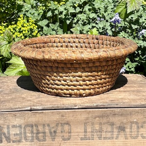 Può includere: Un cesto rotondo e intrecciato con un bordo largo. Il cesto è fatto di fibre colorate naturali, con una fascia più scura attorno al bordo. È appoggiato su una superficie di legno, con vegetazione sullo sfondo. Un oggetto rustico e fatto a mano.