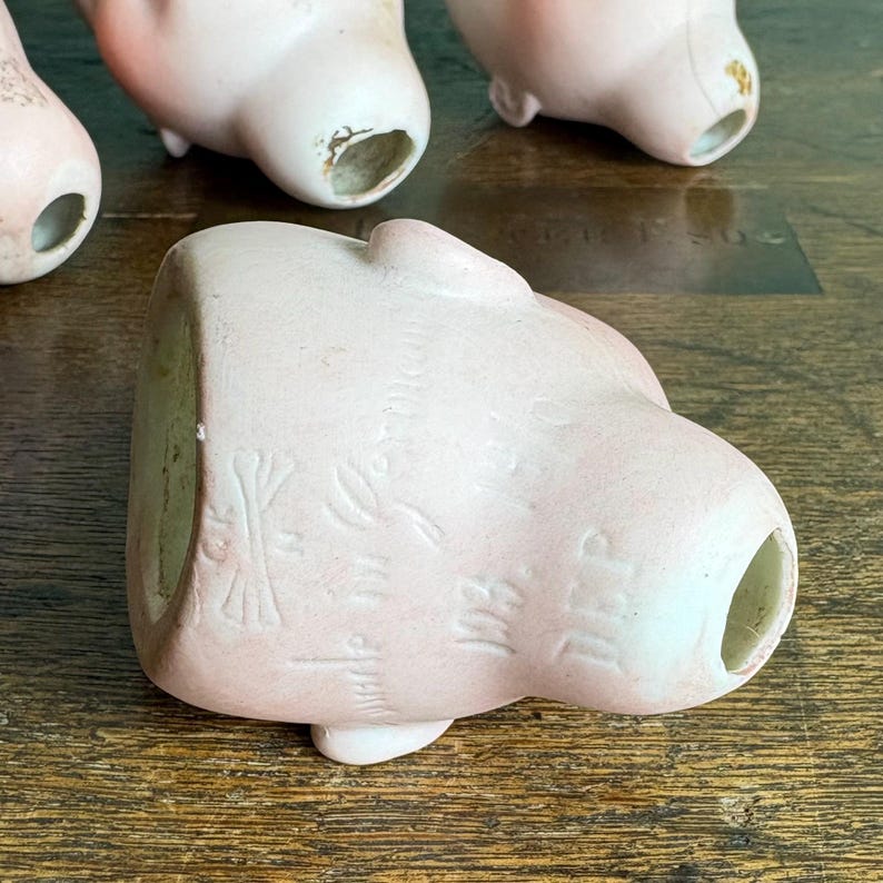 May include: Several antique porcelain doll heads in various stages of wear. The heads are a pale pink color with visible facial features and open neck holes. Some show signs of age and wear, with chipped paint and discoloration.