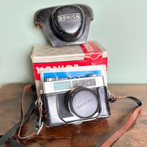 May include: Vintage Konica EE-Matic camera with a black leather case, lens cap, and strap. The camera is silver and black, with the Konica logo visible on the lens cap and camera body. A box and camera cover are also present.