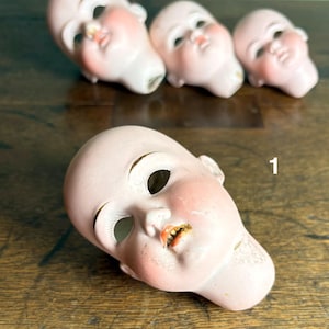 May include: Four antique doll heads with porcelain skin and painted facial features. The heads are a pale pink with rosy cheeks, open mouths, and large eye sockets. The number "1" is visible on the wooden surface.