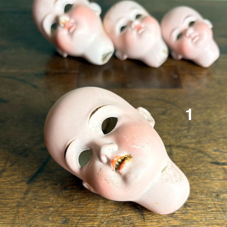 May include: Four antique doll heads with porcelain skin and painted facial features. The heads are a pale pink with rosy cheeks, open mouths, and large eye sockets. The number "1" is visible on the wooden surface.