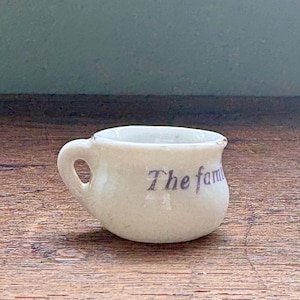 Könnte beinhalten: Eine kleine, cremefarbene Keramiktasse mit Henkel. Auf der Seite der Tasse steht in Dunkelblau "The fam". Die Tasse steht auf einer Holzoberfläche.