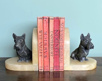 May include: A pair of alabaster bookends featuring a sculpted terrier dog on each side. The bookends are holding up a set of six red books with titles including "Wye Valley", "Warwick", "Canterbury", "London", and "Stratford-upon-Avon".