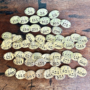 May include: A collection of small, oval-shaped brass tags, each featuring the letters JB and a unique identification number stamped in black. The tags are arranged in rows, with each piece containing two small holes for attachment purposes.