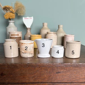 May include: Ten small ceramic jars and a white ceramic measuring cup with the text "TABLE CM" and the number "3" on it. The jars are in various shades of beige and brown.