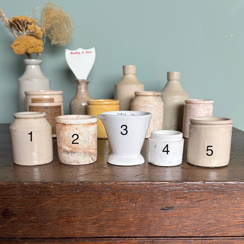 May include: Ten small ceramic jars and a white ceramic measuring cup with the text "TABLE CM" and the number "3" on it. The jars are in various shades of beige and brown.