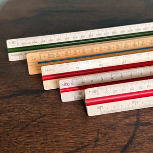 May include: A collection of four triangular architectural scale rulers featuring white faces with black numerical markings and colored grooves in dark green, blue, and two shades of red. Each ruler displays specific ratios like 1:10, 1:100, 1:400, and 1:1250.