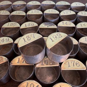 Può includere: Una collezione di contenitori in metallo antico con coperchi in ottone, ciascuno contrassegnato da un numero univoco. I contenitori sono cilindrici con un coperchio a semicerchio e i numeri sono stampati in un carattere classico. Il metallo ha un aspetto invecchiato.