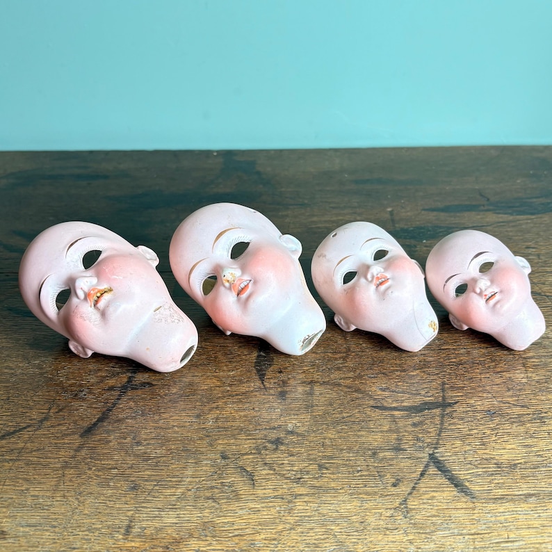 May include: Four antique doll heads with painted features, including rosy cheeks, open mouths, and detailed eyes. The porcelain heads are in varying sizes and rest on a wooden surface, showcasing their delicate craftsmanship and vintage aesthetic.