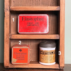 May include: A wooden shelf displays vintage first aid supplies. The shelf holds a red Elastoplast tin of first aid dressings, a smaller Elastoplast tin of elastic adhesive bandages, and a white Mentholatum jar with a metal lid.