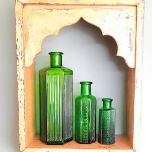 May include: Three green glass bottles with ribbed designs are displayed on a shelf in a wooden frame. The bottles are of different sizes and have a vintage look.