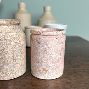 May include: Three white ceramic jars with a crackled glaze finish. The jars are different sizes and shapes. The jars are sitting on a wooden surface.