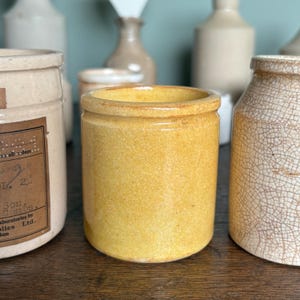 May include: A yellow ceramic jar with a wide mouth and a smooth, slightly textured surface. The jar is sitting on a wooden surface.