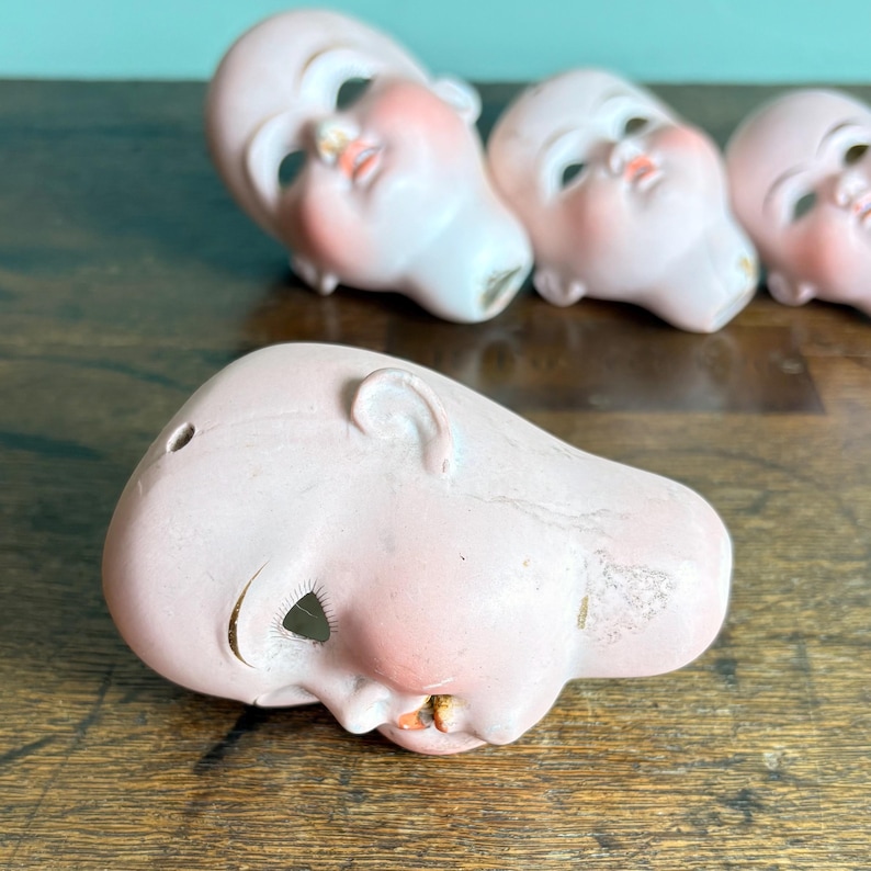 May include: Several antique doll heads are arranged on a wooden surface. The porcelain heads are painted with delicate features, including rosy cheeks, closed eyes, and small mouths. The heads are a pale pink color, with some showing signs of age and wear.