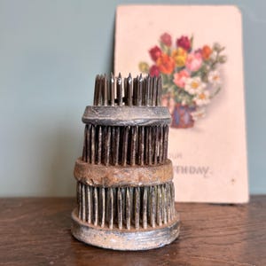 Op de afbeelding: Een vintage bloemenkikker met drie niveaus en metalen pinnen. De kikker is gemaakt van verouderd metaal en heeft een verweerde uitstraling. Een bloemenpostkaart staat op de achtergrond.