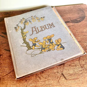 Może przedstawiać: Album w stylu vintage z jasnoszarą okładką z napisem "Album" w złotych literach. Okładka przedstawia fantazyjną ilustrację postaci z kreskówek w niebieskich strojach kąpielowych i żółtych uszach bawiących się pod drzewem.