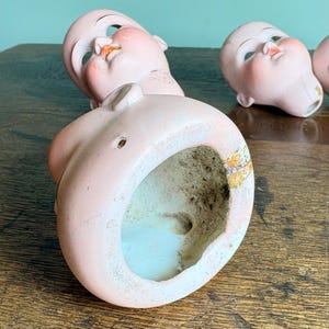 May include: Several antique doll heads with painted facial features, including rosy cheeks and red lips. The porcelain heads are a pale pink color and show signs of age and wear. The heads are resting on a wooden surface.