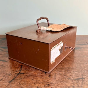 May include: A vintage brown metal box with a handle and keyhole. The box has a small, ornate handle and a key inserted in the lock. A paper tag is attached to the handle. The box is likely used for storage or as a small safe.