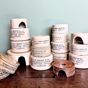 May include: A selection of vintage, cream-coloured ceramic containers and lids, some stacked, with black text. The containers are cylindrical, some with arched openings. The text includes company names and addresses, and the words "London" and "England".