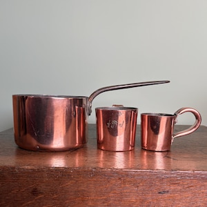 May include: A copper saucepan with a long handle and two copper measuring cups. The measuring cup in the middle has the text "½ Pint" engraved on it. The items are on a wooden surface.