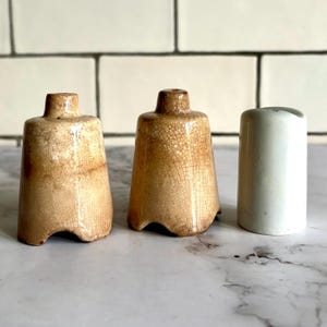 Op de afbeelding: Drie keramische zout- en peperstrooiers. Twee zijn bruin met een gekrakelde glazuur en één is wit.