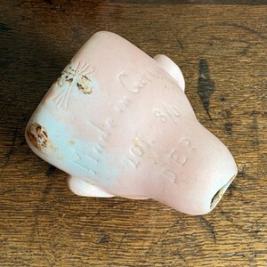 May include: A vintage doll head in a soft pink hue, with visible text including "Made in Germany" and "DEP". The porcelain head has a slightly weathered appearance with areas of light blue paint and brown discoloration, suggesting age and use.