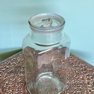 Może przedstawiać: Szklany słoik z pokrywką. Słoik jest pusty i ma kwadratowy kształt. Pokrywka jest biała i ma mały guzik na górze.