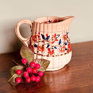 May include: A vintage, cream-colored pitcher with a floral design in red, blue, and gold. The pitcher has a ribbed texture and a curved handle. A sprig of red berries and green leaves rests in front of the pitcher.
