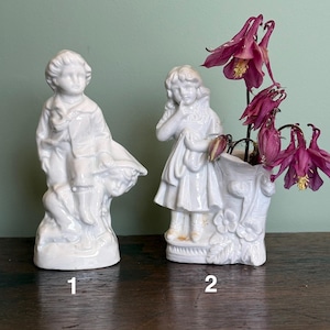 Può includere: Due statuette di porcellana bianca. Una raffigura un ragazzo con un cesto, l'altra una ragazza con un vestito, che tiene un panno. La statuetta della ragazza è un vaso con fiori rosa scuro. Le statuette sono numerate 1 e 2.
