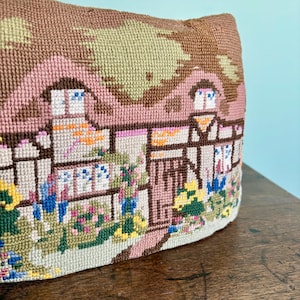 Può includere: Un cuscino arazzo con una scena dettagliata di un cottage con fiori e vegetazione. Il cuscino ha una combinazione di colori marrone, rosa e verde. Il disegno a punto ago raffigura una pittoresca scena di villaggio con dettagli intricati.