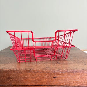 May include: A vibrant red wire desk tidy with a rectangular form. The organiser features a raised central section and is constructed from thin, closely spaced wires. It rests on a wooden surface.
