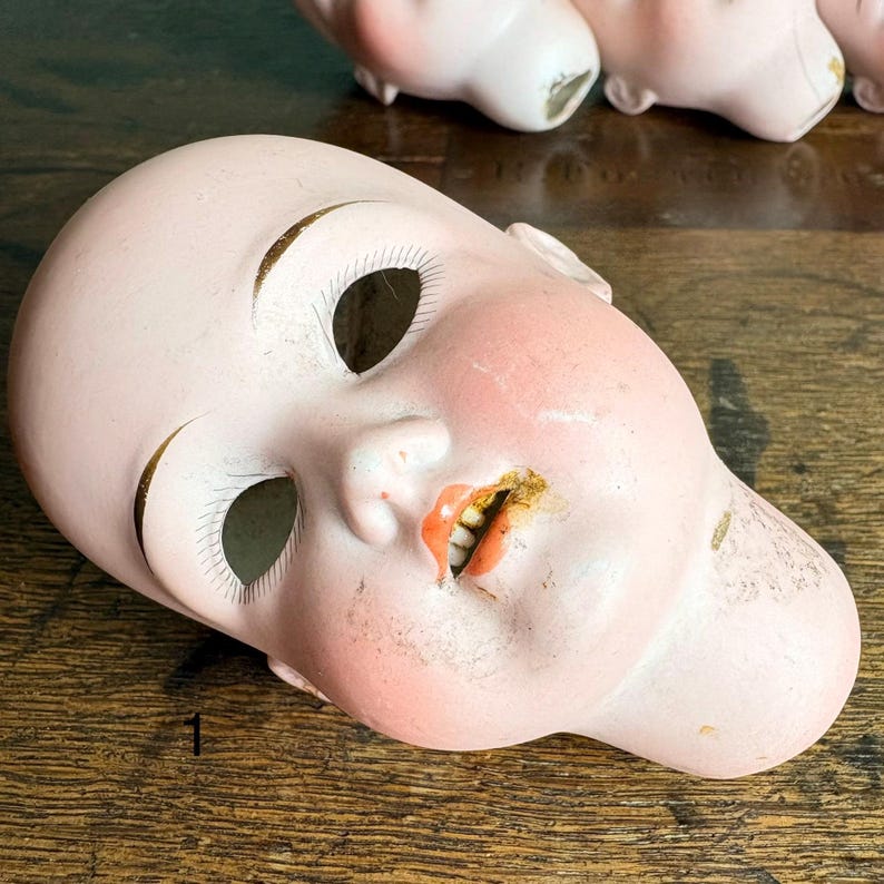 May include: A vintage doll head with a pale pink complexion, open eye sockets, and a partially open mouth revealing teeth. The doll head has painted features, including blush on the cheeks and gold accents around the eyes. The head is lying on a wooden surface.