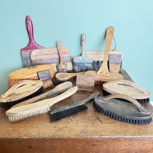 May include: A collection of various vintage brushes with wooden handles and natural or synthetic bristles. The assortment includes flat paint brushes, oval scrubbing brushes, and handheld utility brushes with worn textures and varied wood tones.